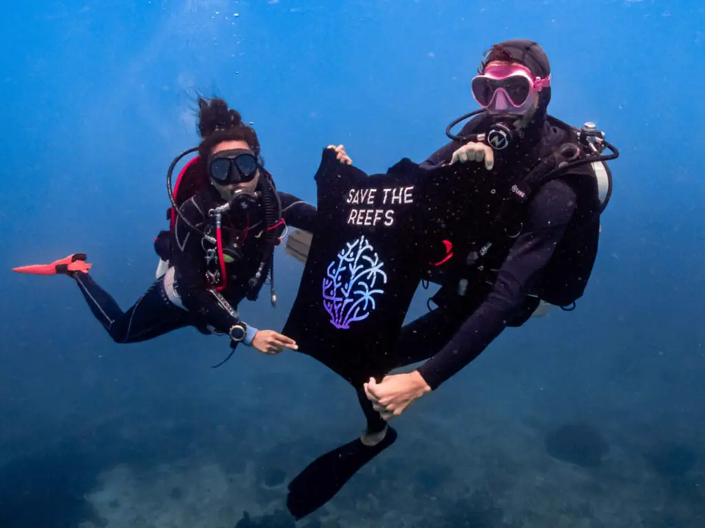 Two divers holding up a Save The Reefs t-shirt underwater in Bira